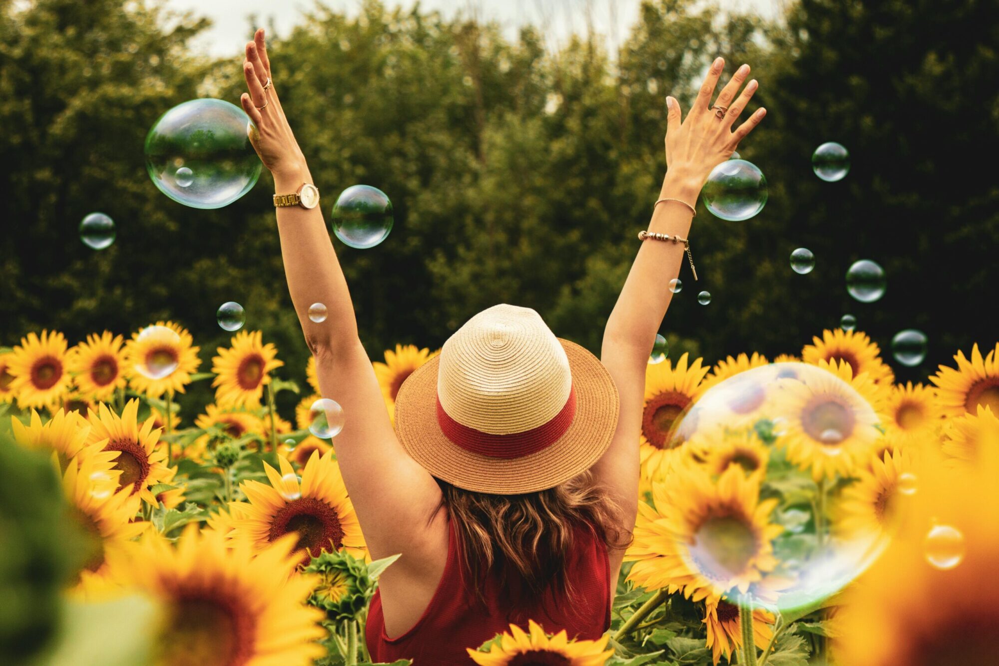 A woman raising her hands to the sky in a field of sunflowers with bubbles floating in the air