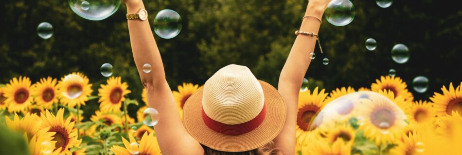 A woman raising her hands to the sky in a field of sunflowers with bubbles floating in the air