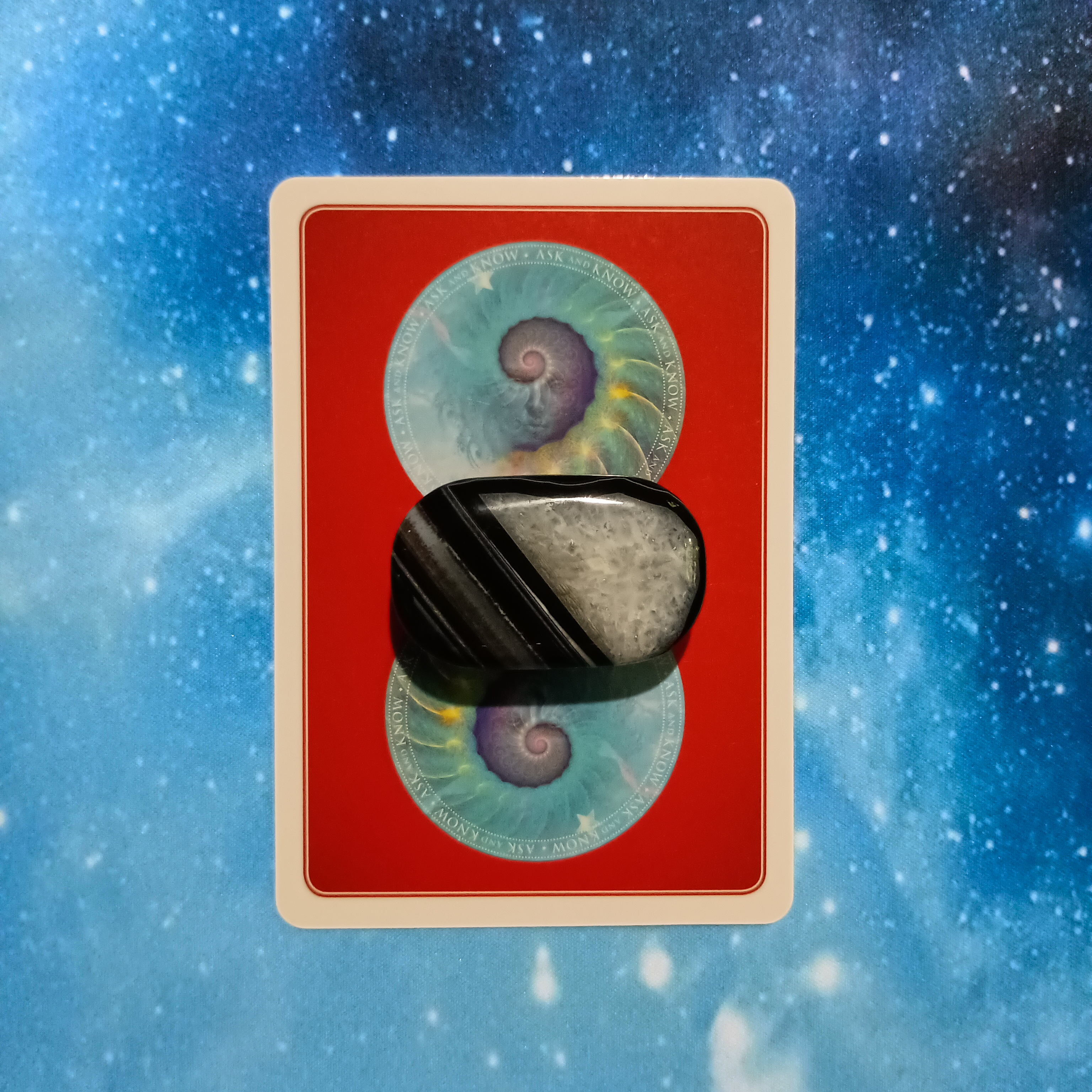 New Moon Pick-a-Card Tarot Reading for May 2025, pile 2 - Black Agate stone and an oracle card on a starry blue background