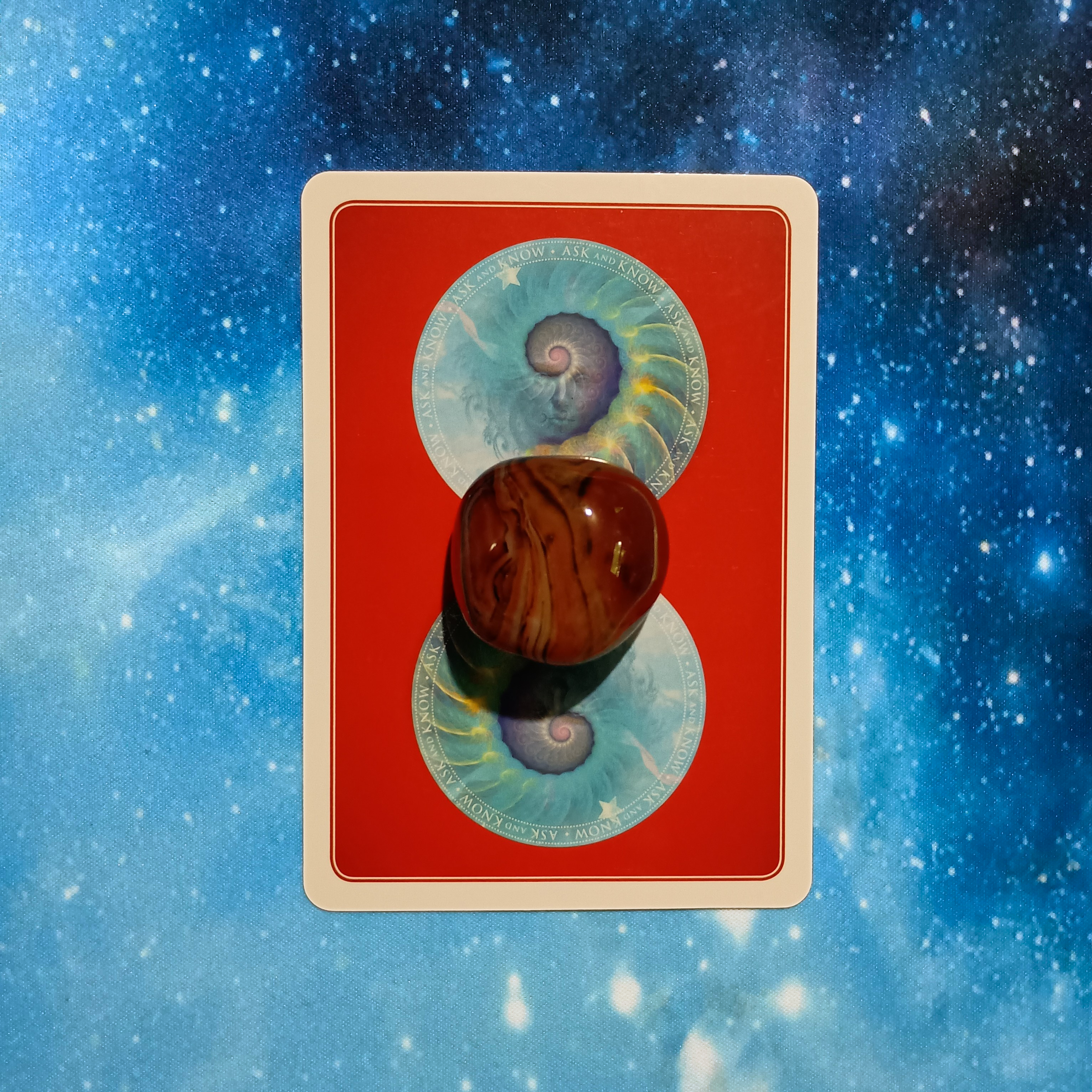 New Moon Pick-a-Card Tarot Reading for May 2025, pile 4 - Silk Agate (red and brown) stone and an oracle card on a starry blue background