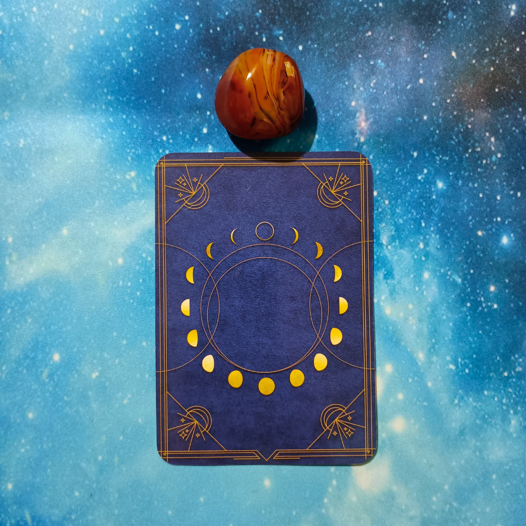New Moon Magic: Your August 2025 Pick-a-Card Tarot Reading, pile 2 — silk agate (orange and brown) stone and an oracle, face down on a starry blue background